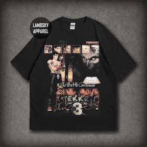 Op de afbeelding: Een zwart t-shirt met korte mouwen en een grafische print van het videospel Tekken 3. Het ontwerp bevat karakterportretten, het Namco-logo, de tekst The Battle Continues en het grote gestileerde Tekken 3-logo in rood en wit.