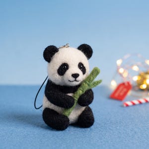 May include: A needle-felted panda bear figurine with black and white wool, featuring black eye patches, ears, and limbs. The panda is holding a small, textured green felt bamboo stalk in its front paws and includes a black cord loop for hanging.