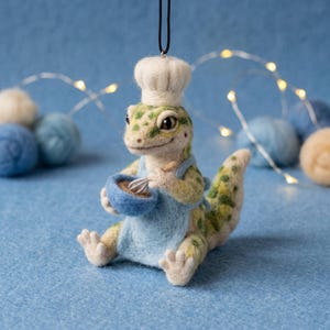 May include: A needle-felted gecko ornament wearing a white chef hat and a light blue apron. The gecko is holding a small blue bowl and a tiny whisk, with green and yellow spotted skin, suspended by a black string for hanging as a decoration.