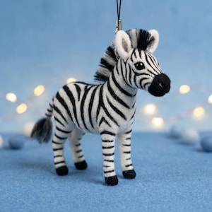 May include: A needle-felted zebra ornament featuring a white body with black stripes, a black mane, a black tail tuft, and a black nose. The zebra has a black string loop attached to the top of its head for hanging, with a small metal ring connector.
