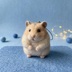 Könnte beinhalten: Eine nadelgefilzte Hamsterfigur mit hellbraunem und weißem Fell, kleinen dunklen Augen und einer rosa Nase. Der Hamster hat einen runden Körper, winzige Pfoten und eine schwarze Kordelschlaufe, die oben am Kopf als Anhänger befestigt ist.