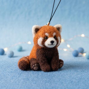 May include: A needle-felted red panda figurine with a soft, fuzzy texture. The creature features a rust-orange back and head, dark brown chest and limbs, and a white face mask. It has a black nose, small dark eyes, and a black cord loop attached.