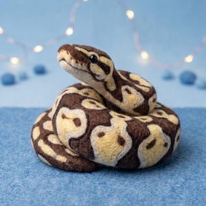 Puede incluir: Una figura de serpiente de lana fieltrada con aguja que presenta un cuerpo enroscado con un patrón de color marrón oscuro y crema. La serpiente tiene una superficie texturizada, la cabeza levantada y pequeños ojos de cuentas negras.