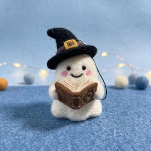 Puede incluir: Una figura de fantasma blanco de lana fieltrada que lleva un sombrero de bruja negro con una hebilla amarilla. El fantasma tiene ojos negros pequeños, mejillas rosadas, una boca sonriente y sostiene un libro marrón abierto.