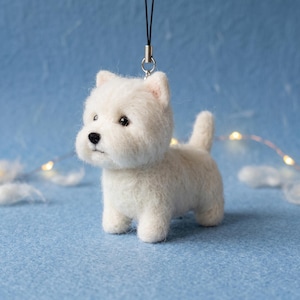 Puede incluir: Un adorno de perro West Highland White Terrier de lana fieltrada blanca con textura difusa, ojos de cuentas negras, una pequeña nariz negra y un cordón negro con un accesorio de metal plateado para colgar como dije.
