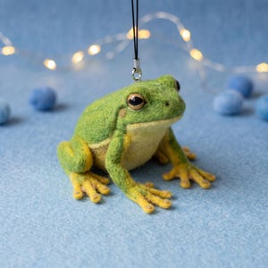 May include: A needle-felted frog ornament featuring a vibrant green back, a pale yellow underside, and detailed webbed feet. The frog has dark, expressive eyes and is attached to a thin black hanging cord with a silver metal loop.