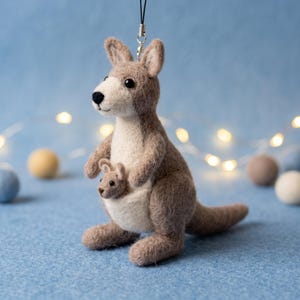 Könnte beinhalten: Ein aus Filzwolle gefertigtes Känguru-Ornament mit hellbraunem Körper sowie cremefarbener Brust und Schnauze. Ein kleines Jungtier befindet sich im Beutel. Die Figur hat schwarze Perlenaugen, eine schwarze Nase und eine schwarze Aufhängeschlaufe.