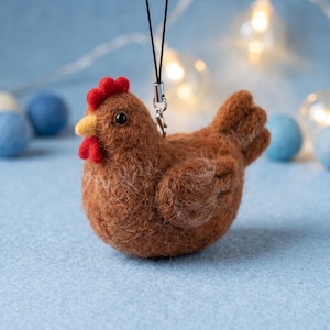 May include: A brown needle-felted chicken ornament with a bright red comb and wattle, a yellow beak, and black bead eyes. The chicken is attached to a black cord with a silver-toned metal clasp for hanging, featuring a textured wool finish.