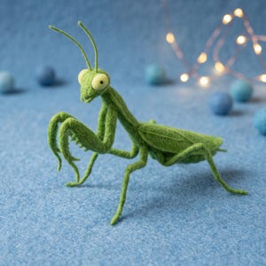 May include: A needle-felted green praying mantis figurine with two long antennae, large pale yellow eyes, and articulated legs. The insect sculpture features a textured fiber surface and is posed in a standing position with its front raptorial legs folded.