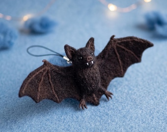 Op de afbeelding: Een donkerbruine, naaldgevilte vleermuis-ornament met een textuurrijk wollen lichaam, puntige oren en twee kleine witte hoektanden. De vleermuis heeft uitgestrekte vleugels en een zwart koordje aan de bovenkant om als decoratieve hanger te dienen.