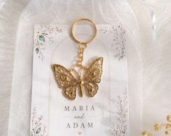 Puede incluir: Un llavero de mariposa de metal dorado sujeto a una tarjeta rectangular con ilustraciones florales y el texto impreso MARIA and ADAM Wedding Favour, todo dentro de una bolsa de organza blanca transparente.
