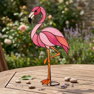 Puede incluir: Una escultura de flamenco hecha de vidrieras con paneles de vidrio en varios tonos de rosa y coral unidos por líneas de metal oscuro. El ave se sostiene sobre una pata de metal naranja con una base palmeada, mostrando un cuello curvado.