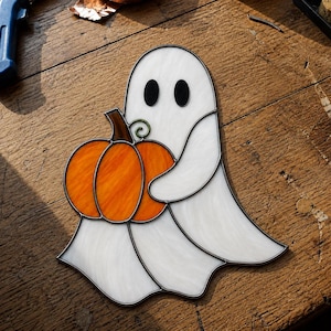 May include: A stained glass suncatcher shaped like a white ghost with two black oval eyes, holding a bright orange pumpkin with a brown stem and a small green vine detail. The piece is constructed with dark metal solder lines between glass segments.
