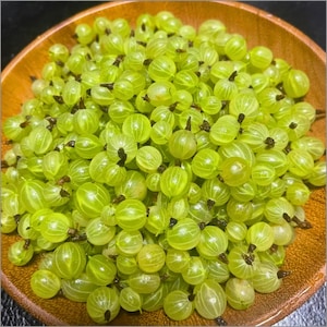 Puede incluir: Una gran cantidad de grosellas espinosas frescas, translúcidas y de color verde claro, amontonadas en un cuenco de madera redondo. Cada fruta pequeña y redonda tiene una piel lisa y veteada con un pequeño resto de flor marrón oscuro.