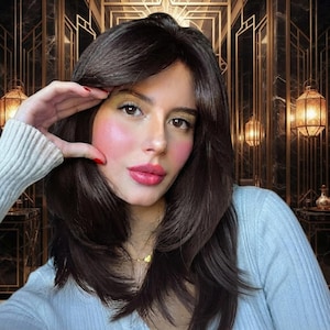 May include: A person with dark brown hair styled in a layered cut with curtain bangs, wearing a light blue ribbed long-sleeve top. The person has bright pink blush on their cheeks and red nail polish on their fingernails, with a gold necklace.