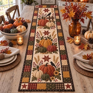 Puede incluir: Un camino de mesa rectangular de tela con un diseño acolchado que presenta tres paneles centrales con calabazas, espigas de trigo, bayas, girasoles y pájaros negros. El borde consiste en un patrón de patchwork de cuadrados en tonos otoñales.