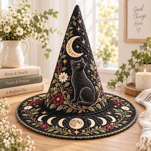 May include: A black quilted witch hat featuring an embroidered design of a sitting black cat, a crescent moon, stars, and floral motifs. The wide brim displays a lunar cycle phase pattern with red flowers and green leaves stitched throughout.