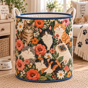 May include: A cylindrical fabric storage basket featuring a quilted pattern of various cats and colourful flowers. The basket has a dark blue rim, white interior lining, and two cream-coloured fabric handles attached to the top sides.