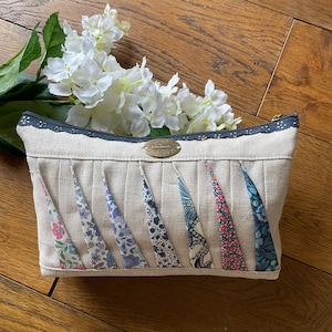 Könnte beinhalten: Eine beige Kosmetiktasche mit Reißverschluss und dekorativer Spitzenborte. Die Tasche hat vertikale Falten und farbenfrohe Stoffakzente mit floralen Mustern. Ein kleines Metallschild ist vorne angebracht.