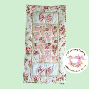 Op de afbeelding: Een roze en groene quilt van bloemenstof met een hartvormig ontwerp en een strik aan de boven- en onderkant. De quilt is omlijst met een groene rand. De tekst "PDF Sewing Pattern Sallieann Harrison - Quilter" staat op een witte cirkel met een roze naaimachine.