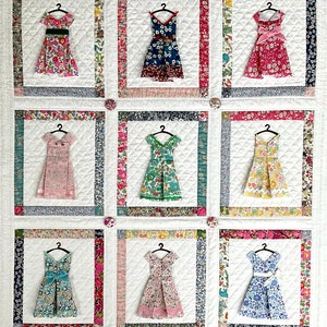 May include: A white quilt with nine framed squares, each featuring a different patterned dress on a hanger. The dresses are in various colours and patterns, including floral, polka dot, and geometric.