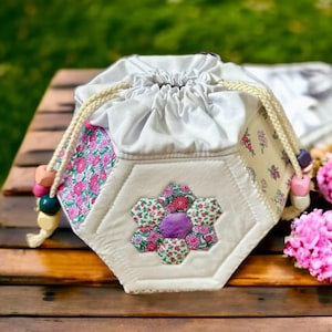 May include: A handcrafted drawstring bag with a floral patchwork design. The bag has a white body with a flower applique and pink and green floral fabric accents. The beige drawstring has wooden beads.