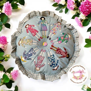 May include: A round cushion with a blue background and a white gingham trim.  The cushion is decorated with 12 embroidered figures of girls wearing floral dresses.  The text "PDF Sewing Pattern" and "Sallieann Harrison - Quilter" is printed on a white circle with a pink sewing machine.