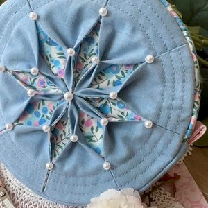 May include: A round, light blue fabric hat featuring a starburst pattern. The star is crafted from floral fabric and embellished with faux pearls. The hat's brim is a solid light blue. The design has a vintage, handmade feel.