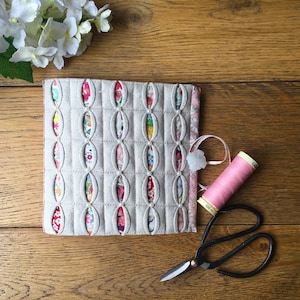 Könnte beinhalten: Eine beige Stoffnadelbox mit einem gesteppten Design mit ovalen Ausschnitten. Die Box hat einen rosa Stoffbesatz und ein weißes Band mit einem weißen Knopf. Eine rosa Garnrolle und eine schwarze Schere sind ebenfalls abgebildet.