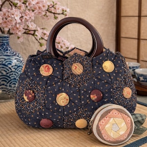 May include: A navy blue quilted handbag with a wooden handle and decorative floral buttons. The bag features a geometric pattern and is accompanied by a small, round coin purse with a similar design. The text "Quilt the Life" is visible on the bag.