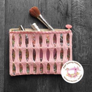 Puede incluir: Bolsa de maquillaje de tela rosa con un diseño recortado y una cremallera blanca. La bolsa está llena de pinceles de maquillaje y botellas. Un círculo blanco con una máquina de coser rosa y flores se encuentra en la esquina inferior derecha. El texto en el círculo dice "PDF Sewing Pattern Sallieann Harrison - Quilter".