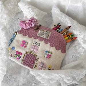 Puede incluir: Un estuche con forma de casa con un techo festoneado rosa y una cremallera. El estuche está decorado con detalles florales bordados, el año "2015" y una pequeña flor. Se ven lápices de colores dentro.