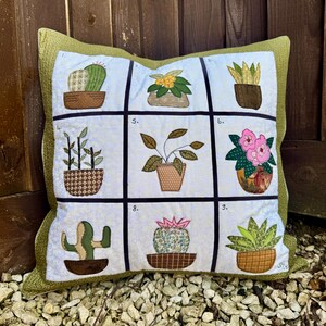 May include: A green and white pillow with nine squares featuring different potted plants. Each square has a number in the top corner. The plants include cacti, succulents, ferns, and flowering plants.