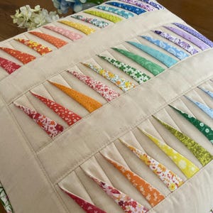 May include: A square, beige pillow with a quilted design. The pillow features rows of colorful, patterned fabric strips in a rainbow of colors, including red, orange, yellow, green, blue, and purple. The strips are arranged in a fan-like pattern.
