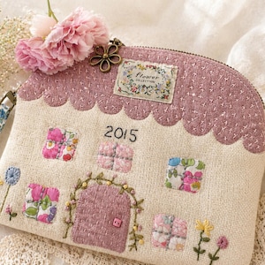 Op de afbeelding: Een handgemaakte, huisvormige etui met een roze dak en deur, met bloemenstof ramen en geborduurde details. De etui bevat de tekst "2015" en een bloemenlabel. Een roze bloem en een bloemenbedel zijn bevestigd.