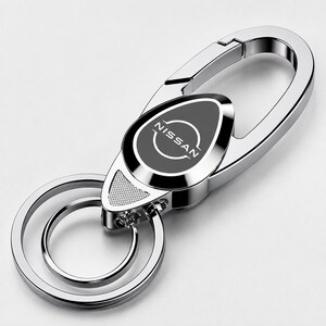 May include: A silver-toned metal keychain featuring a polished carabiner clip and two circular rings. The central oval emblem is black with the white Nissan logo and text printed in the center. The design includes a small textured triangular detail.