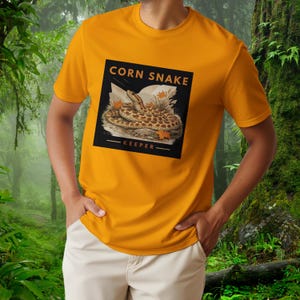 Könnte beinhalten: Ein senfgelbes T-Shirt mit einem quadratischen Grafikdruck auf der Brust. Das Design zeigt eine zusammengerollte Kornnatter auf einem felsigen Untergrund mit Herbstblättern und dem Text CORN SNAKE KEEPER in einer dunklen Schrift.
