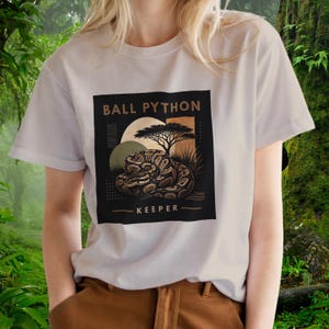 Puede incluir: Una camiseta de color claro con un gráfico cuadrado negro en el pecho. El diseño muestra una pitón bola enroscada, la silueta de un árbol y formas circulares en tonos beige y verde oliva. El texto dice BALL PYTHON KEEPER en letra sans-serif.
