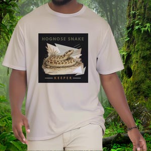 以下が含まれることがあります： 胸元に四角いグラフィックがプリントされた白い半袖Tシャツ。グラフィックには、砂地の上にいる茶色のホグノーズスネークが描かれており、幾何学模様と枯れ草を背景に「HOGNOSE SNAKE KEEPER」という文字が記されています。