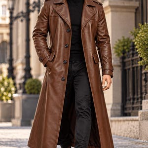 May include: A long brown leather trench coat featuring a wide collar, shoulder epaulets, and four front buttons. The coat is worn over a black turtleneck sweater and black trousers, paired with brown leather boots and dark sunglasses.