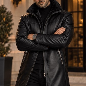 May include: A black leather mid-length coat featuring a plush black faux fur collar, a full-length front zipper, and two side zippered pockets. The coat is worn over a black turtleneck sweater and black pants, with a silver wristwatch visible.