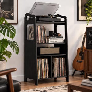 May include: A black wooden record player stand featuring a top shelf for a turntable, a middle shelf with books titled The Vinyl Book, Rolling Stones, and A Life in Music, and two lower compartments filled with vinyl records in sleeves.