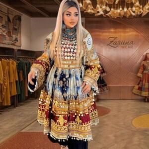May include: A long-sleeved dress featuring a vibrant mix of blue, white, and deep red patterns, heavily embellished with gold-toned coin accents, intricate embroidery, and metallic trim. The garment includes a high neckline and flared sleeves.