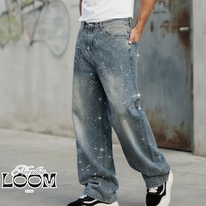 May include: Blue denim jeans featuring a white paint splatter pattern, a high-waisted fit with belt loops, five pockets, and a wide-leg silhouette. The jeans have a faded wash effect and are paired with a plain white t-shirt and black trainers.