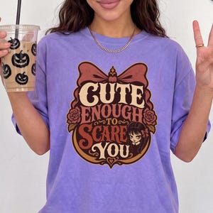 Peut inclure: Un t-shirt violet clair avec un motif graphique comprenant un grand nœud, des roses et le texte "CUTE ENOUGH TO SCARE YOU" dans une police stylisée. Le design inclut une petite illustration d'un personnage aux cheveux foncés avec un nœud.