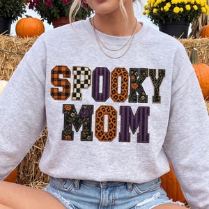 Op de afbeelding: Een lichtgrijs sweatshirt met ronde hals en de tekst SPOOKY MOM in grote blokletters. Elke letter heeft een uniek patroon, waaronder oranje en zwart ruitjespatroon, zwart-wit schaakbordpatroon, paarse strepen, luipaardprint en bloemen.