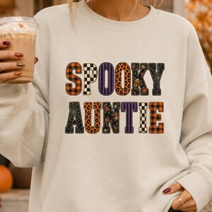 May include: A cream-coloured crewneck sweatshirt featuring the text SPOOKY AUNTIE in large block letters. Each letter displays a different pattern, including orange and black plaid, black and white checkerboard, purple stripes, leopard print, and spiderwebs.