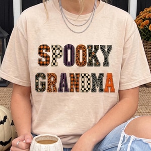 Può includere: Una t-shirt beige a maniche corte con la scritta SPOOKY GRANDMA in stampatello. Ogni lettera presenta un motivo unico, tra cui quadri arancioni, scacchi bianchi e neri, strisce viola, stampa leopardata, fiori e ragnatele.