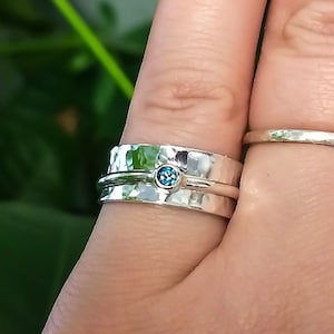 May include: A silver ring with a hammered texture and a small, round, blue gemstone. A second, thinner silver band is also visible. The rings are on a person's finger, with a plain silver band on the adjacent finger. The background is blurred green foliage.
