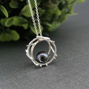 May include: A silver pendant necklace featuring a dark pearl set within a circular, twig-like frame. The pendant hangs from a delicate chain. The design has a natural, organic aesthetic, with a focus on the pearl's luster.
